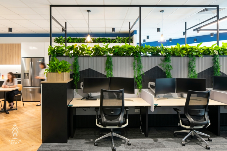 Bright, modern corporate office interior featuring an integrated indoor planter trough by Vertical Green Malaysia. Lush green plants and cascading vines form a natural partition above a row of cubicles with dual-monitor workstations, enhancing biophilic office design.
